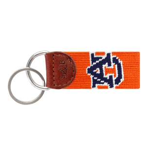 Bright orange needlepoint key fob displaying the navy “AU” Auburn logo, brown leather trim, and a metal key ring.