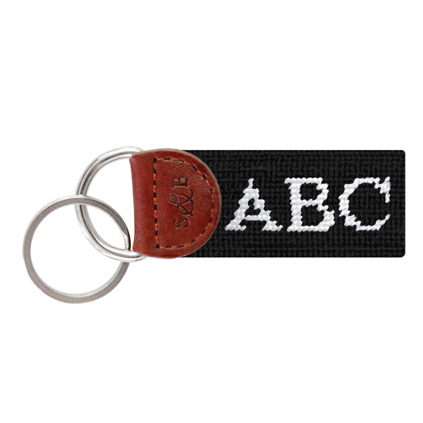 Black needlepoint key fob with white monogram initials, brown leather trim, and a metal key ring.