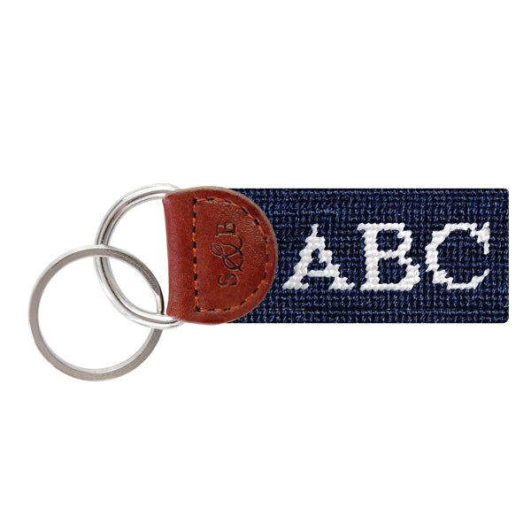 Dark navy needlepoint key fob featuring white monogram initials, brown leather trim, and a metal key ring.