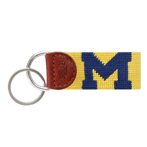 Bright yellow needlepoint key fob featuring the navy block “M” Michigan logo, brown leather trim, and a metal key ring.