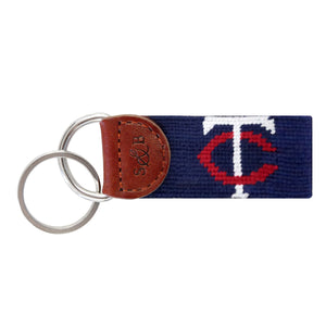 Dark navy needlepoint key fob with the red and white “TC” logo, brown leather trim, and a metal key ring.
