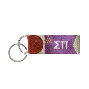 Purple needlepoint key fob displaying the Greek letters ΣΠ in white, brown leather trim, and a metal key ring.