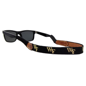 Black needlepoint sunglass strap featuring the Wake Forest logo stitched in gold, with leather backing and cotton wrapped connectors for secure wear