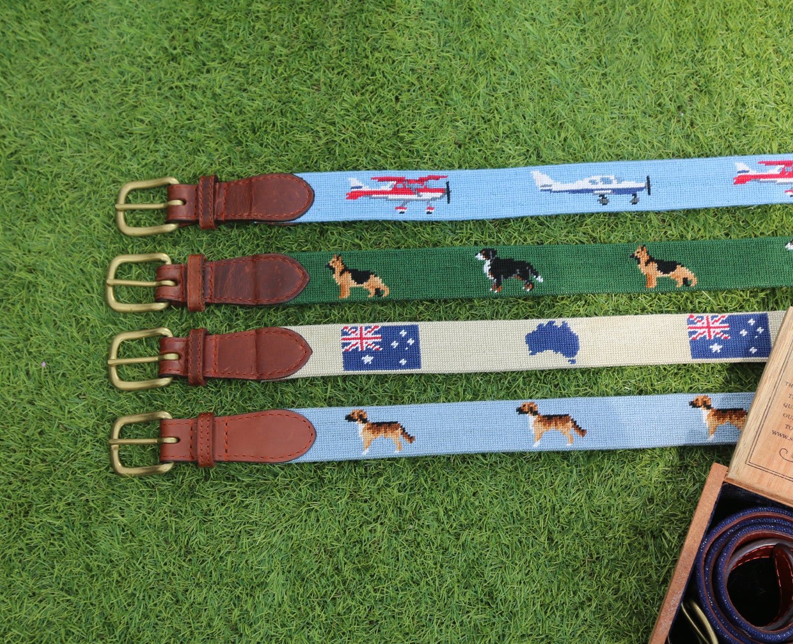 four needlepoint belts laid on grass featuring different motifs: planes, dogs, Australia, dog.