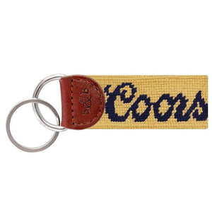 Navy needlepoint key fob featuring the Coors logo with red script and a mountain silhouette, brown leather trim, and a metal key ring.