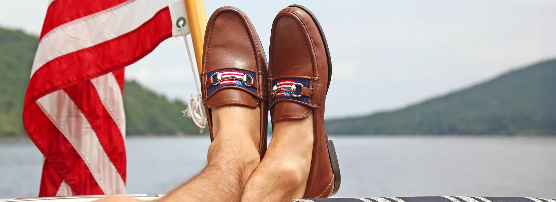 Man's feet posing over scenic lake modeling needlepoint downing bit loafers in American Flag 