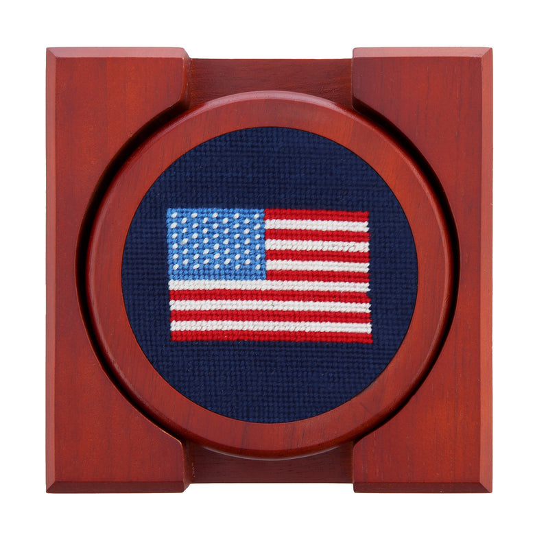 Set of four round wooden coasters with stitched needlepoint logos stacked neatly in a wooden holder.