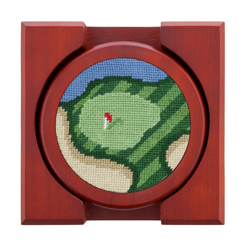 Set of four round wooden coasters with stitched needlepoint logos stacked neatly in a wooden holder.