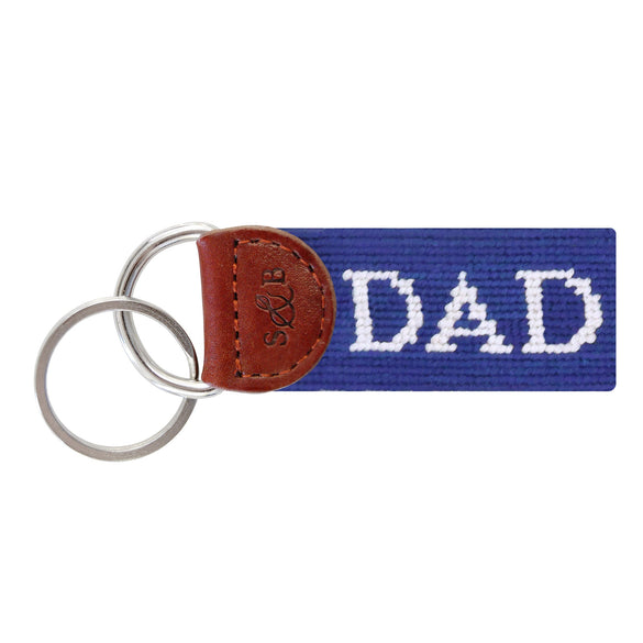 Royal blue needlepoint key fob displaying the word "Dad" in white block letters, brown leather trim, and a metal key ring.