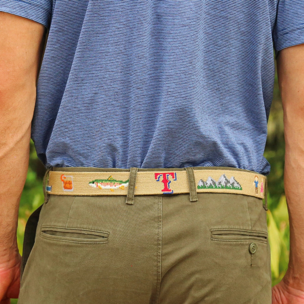 Person wearing a dark khaki needlepoint belt with a colorful design, blue shirt, and green pants against a blurred natural background.