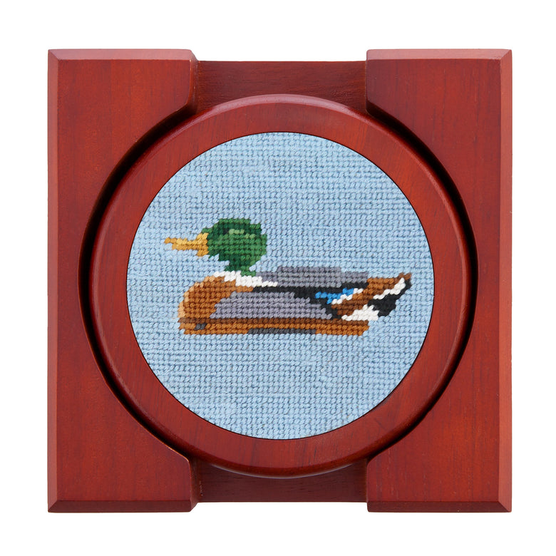 Set of four round wooden coasters with stitched needlepoint logos stacked neatly in a wooden holder.