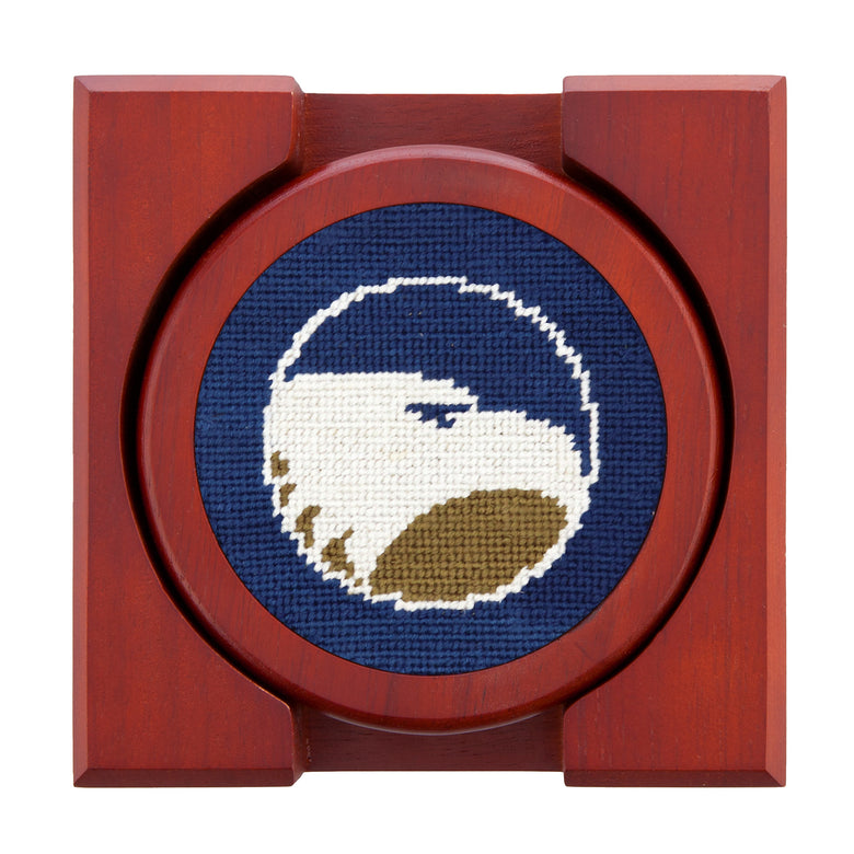 Set of four round wooden coasters with stitched needlepoint logos stacked neatly in a wooden holder.