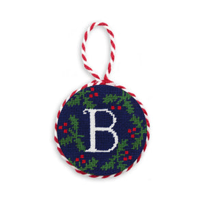 Round needlepoint ornament featuring a white block letter “B” surrounded by holly, with scalloped edges and a red-and-white hanging cord
