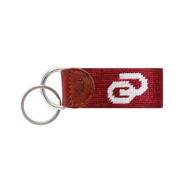 Crimson needlepoint key fob featuring the white Oklahoma “OU” logo, brown leather trim, and a metal key ring.