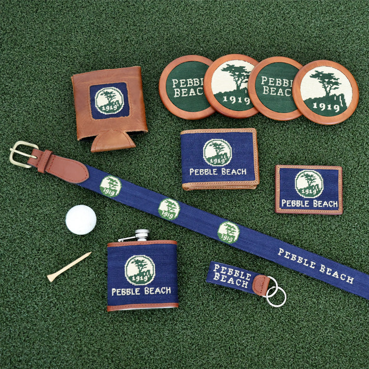 Collection of Pebble Beach branded items including a belt, can cooler, coasters, and keychain on a green background.