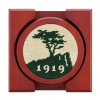 Set of four round wooden coasters with stitched needlepoint logos stacked neatly in a wooden holder.