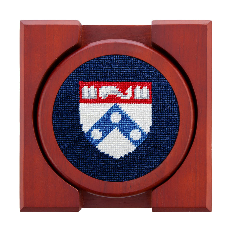 Set of four round wooden coasters with stitched needlepoint logos stacked neatly in a wooden holder.