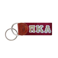 Garnet needlepoint key fob featuring gold and white Pi Kappa Alpha letters, brown leather trim, and a metal key ring.