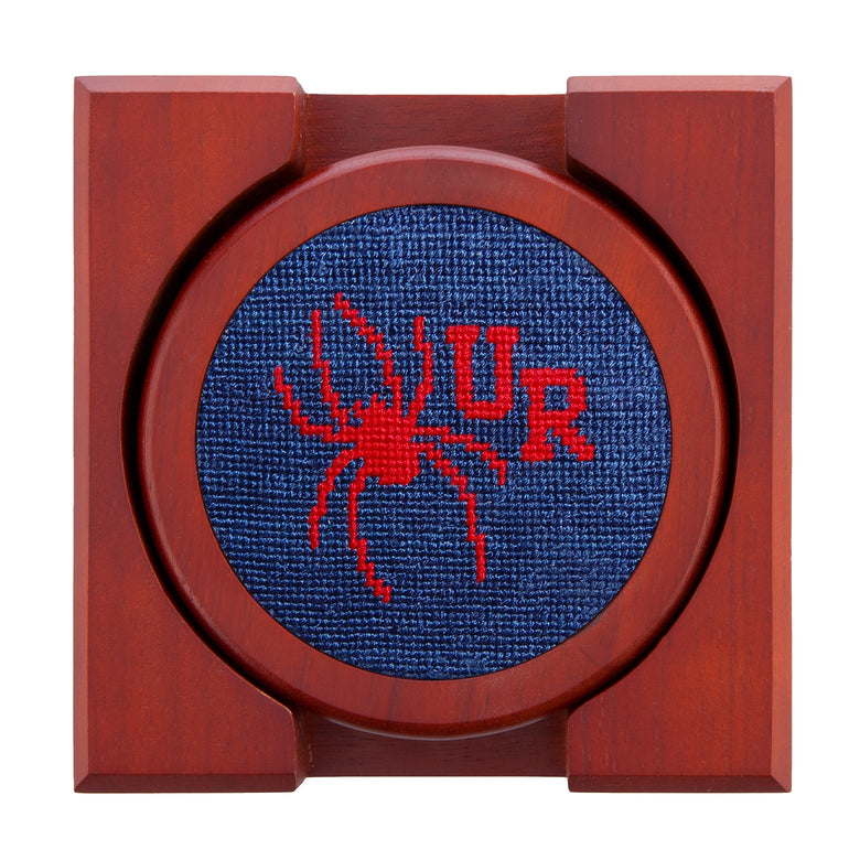 Set of four round wooden coasters with stitched needlepoint logos stacked neatly in a wooden holder.