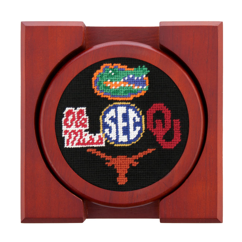 Set of four round wooden coasters with stitched needlepoint logos stacked neatly in a wooden holder.