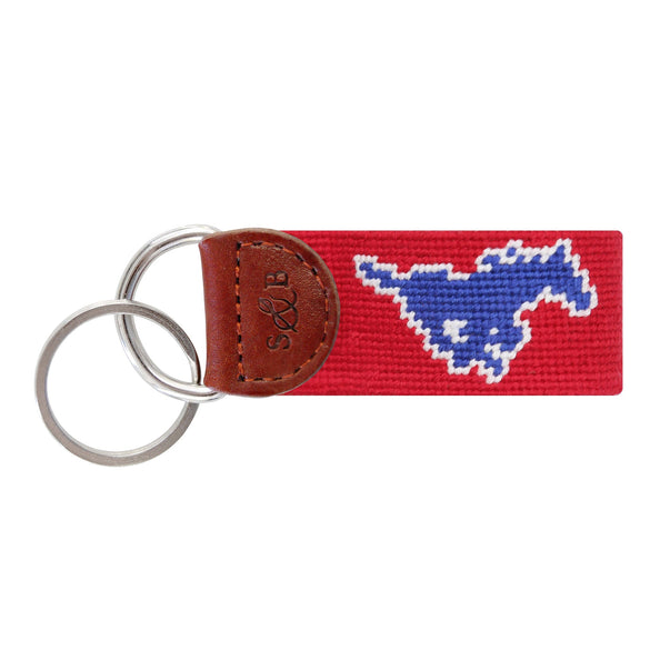 Red needlepoint key fob featuring a blue SMU mustang logo, brown leather trim, and a metal key ring.