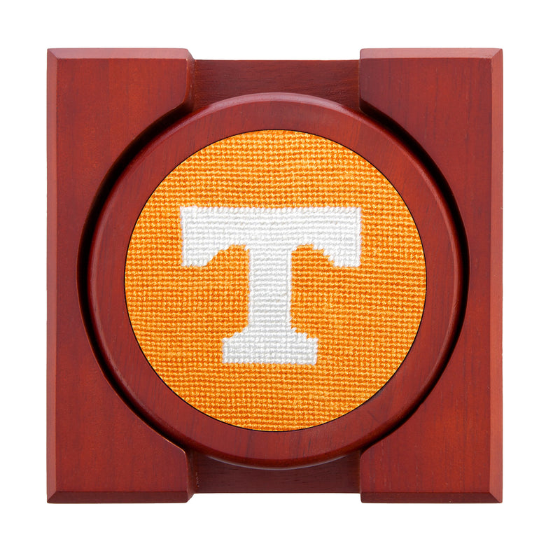 Set of four round wooden coasters with stitched needlepoint logos stacked neatly in a wooden holder.