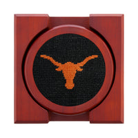 Set of four round wooden coasters with stitched needlepoint logos stacked neatly in a wooden holder.